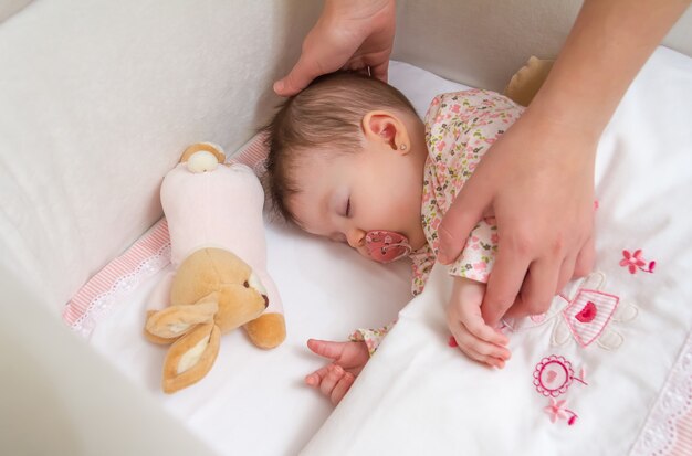 Manos de madre acariciando a su linda niña durmiendo en una cuna con chupete y peluche
