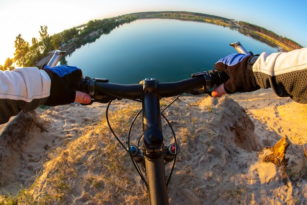 Manos en el manillar de una bicicleta frente al acantilado del lago por la noche. ocio deportivo de verano