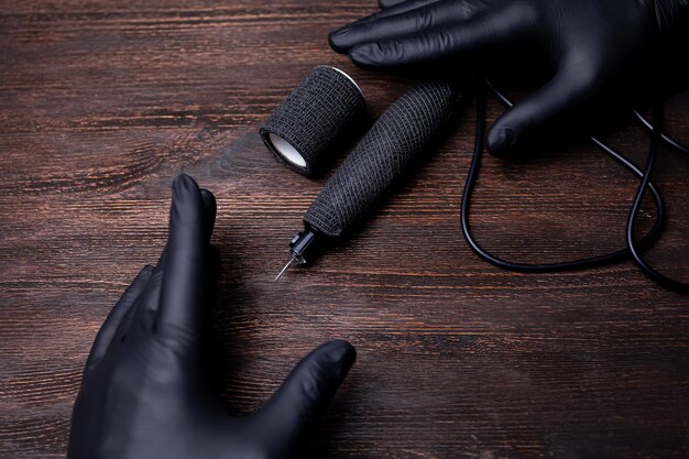 Las manos de un maquillador permanente con guantes estériles negros frente a los cuales se encuentra una máquina de tatuajes envuelta con una cinta de vendaje sobre una mesa de madera