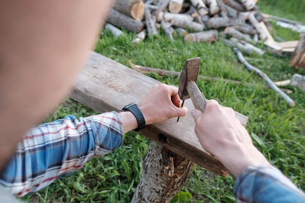 Las manos masculinas de un carpintero hacen un banco martillando clavos en una tabla en el patio de un pueblo