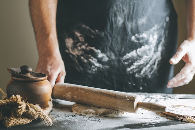 Manos masculinas enrollar la masa y la harina con un rodillo junto a la olla de barro y la botella de aceite en la mesa oscura, mientras se cocina