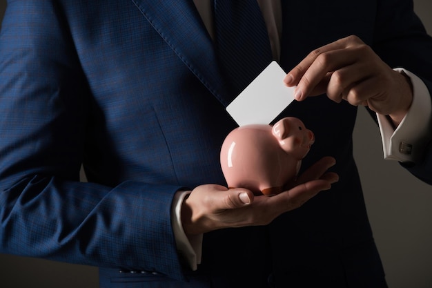 Manos masculinas poniendo la tarjeta de visita en blanco en la hucha para ahorrar sobre fondo gris elegante formal bl ...