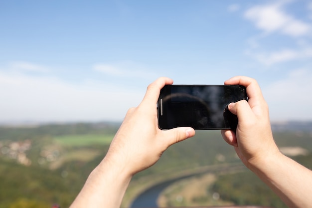 Manos masculinas sosteniendo su teléfono móvil al aire libre, tomando una foto en el parque paisajístico