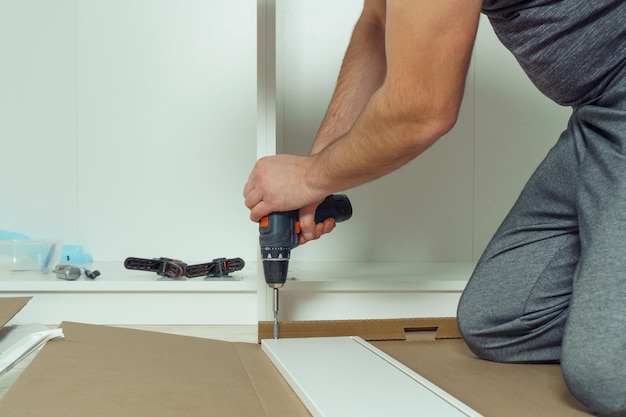 Las manos masculinas sostienen un destornillador eléctrico inalámbrico para fijar las paredes del cajón blanco que ensambla el armario de la cómoda en el piso