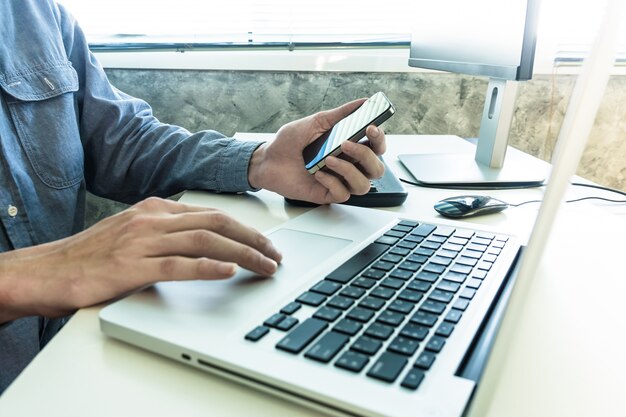 Manos masculinas usando el teléfono inteligente con la computadora en el escritorio.