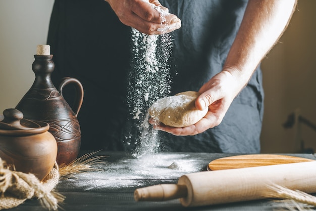 Manos masculinas vierten harina sobre la masa junto a la olla de barro y la botella de aceite y el rodillo sobre la mesa oscura, mientras se cocina