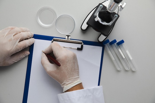 Manos de médico tomando notas en su oficina, tubos de ensayo y microscopio en una mesa