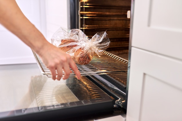 Manos mujer ama de casa cocinando cerdo en casa en una manga para hornear, pone una bandeja de carne en el horno