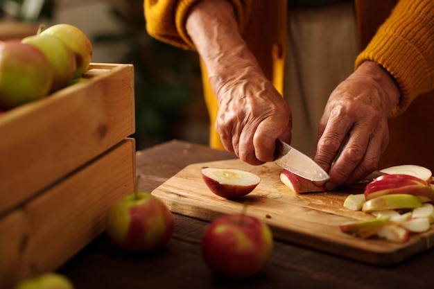 Las manos de una mujer anciana cortando manzanas frescas maduras con un cuchillo en una tabla de madera