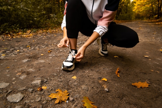 Manos de mujer arreglando los cordones de los zapatos en sus zapatillas. Cerrar vista de corte. Corredor de pista femenino. Concepto de ejecución por la mañana. Joven atlética para correr en el bosque. Concepto de estilo de vida saludable. Follaje de otoño en la carretera.