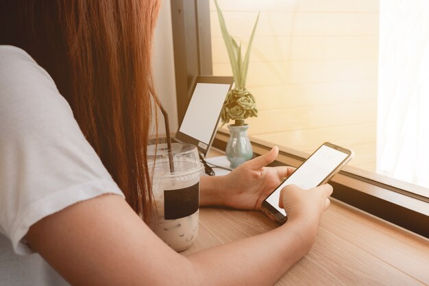 Las manos de la mujer asiática que sostienen el teléfono móvil y la computadora portátil con la pantalla blanca en café café.