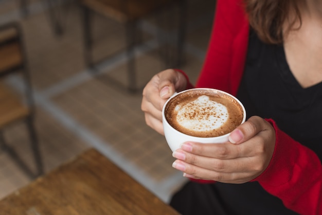 Las manos de la mujer en el cadigan rojo que sostiene la taza de café del cappucino en invierno.