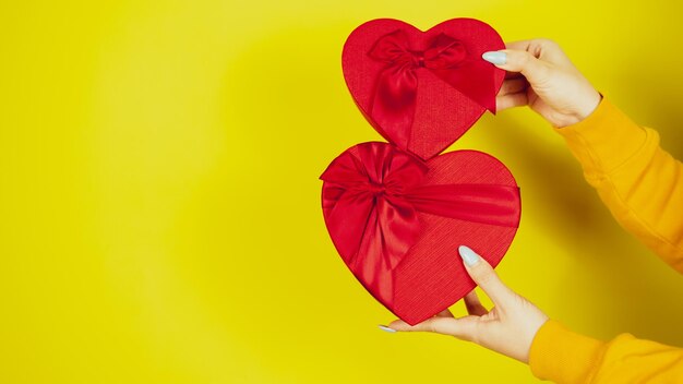 Manos de mujer con cajas de regalo rojas en forma de corazón sobre fondo amarillo Persona irreconocible con regalos románticos con cinta Concepto de presente en el día de San Valentín y el día internacional de la mujer