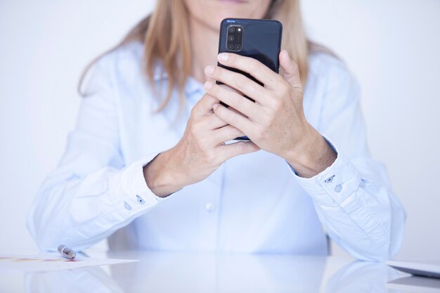 Manos de mujer con una camisa de vestir azul sosteniendo el teléfono móvil con ambas manos mientras trabaja en su oficina y escritorio blanco