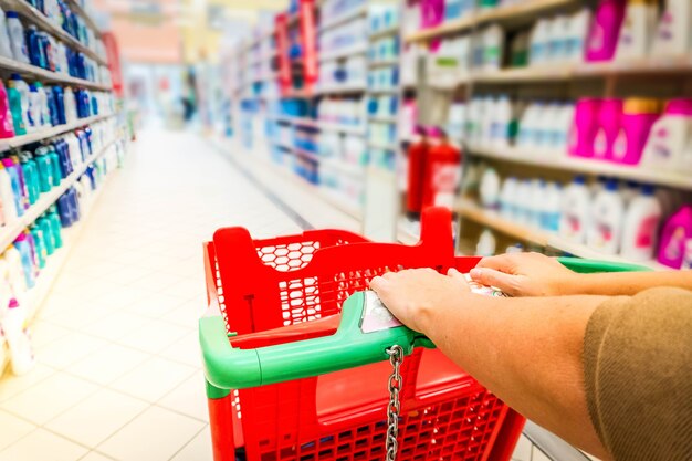 Manos de mujer con carrito de compras de cerca, estantes con mercancías en el fondo, tonificado