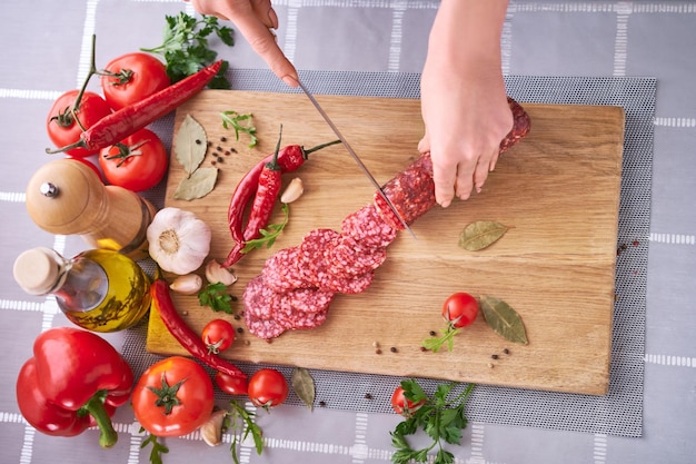 Manos de mujer cortando una salchicha de salami en una tabla de cortar de madera en la cocina doméstica