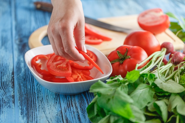 Manos de mujer cortando tomate, detrás de verduras frescas.