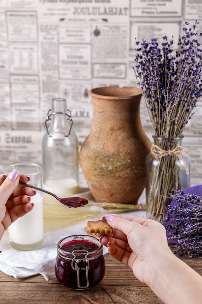 Manos de mujer con cuchara y galleta de miel de azúcar casera. Mermelada de frambuesa en tarro, unch de lavanda sobre un fondo de madera. Concepto de desayuno.