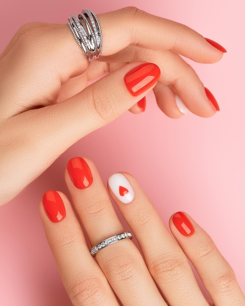 Manos de mujer cuidadas con anillos sobre fondo rosa diseño de uñas rojas de moda con corazón