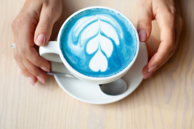 Manos de mujer desenfocada sosteniendo una taza de café con leche de guisante de mariposa caliente o café con leche de espirulina azul en la mesa de madera. Bebida orgánica saludable y de moda. Concepto de bienestar y desintoxicación. Copie el espacio.