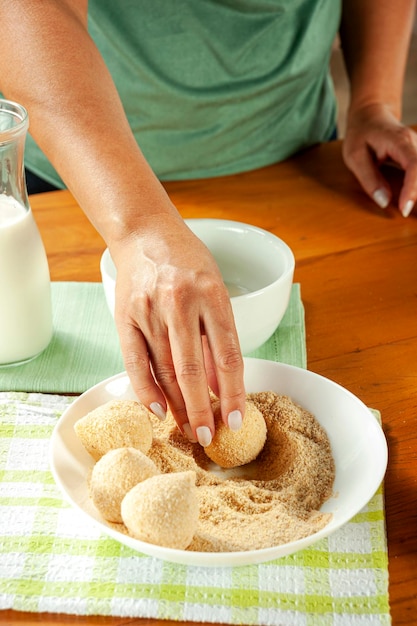 Manos de mujer empanando croqueta brasileña coxinha de frango con pan rallado en una mesa de cocina de madera