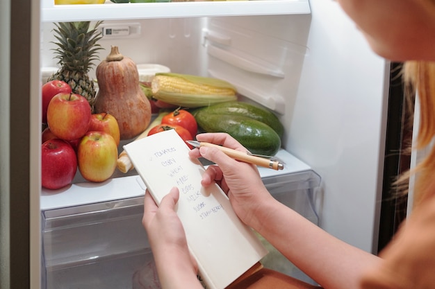 Manos de mujer escribiendo la lista de alimentos que necesita comprar en el supermercado o hacer un pedido en línea