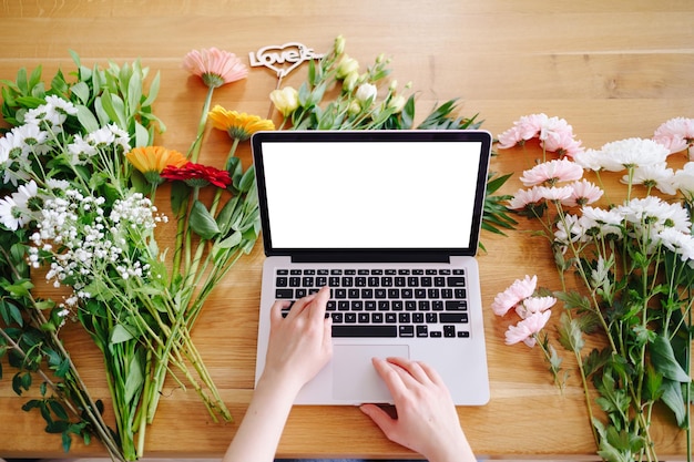 Manos de mujer escribiendo en el teclado de una computadora portátil con pantalla blanca rodeada de coloridas flores frescas y la inscripción amor es el concepto del día de San Valentín