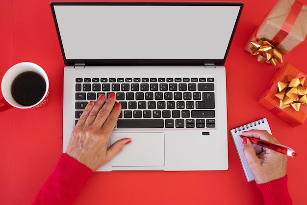 Manos de mujer escribiendo en el teclado de la computadora portátil en el tiempo de Navidad espacio en blanco en la pantalla para texto de fondo rojo con regalos de Navidad