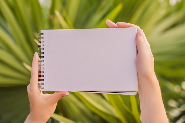 Manos de mujer en un fondo tropical sosteniendo una maqueta de papel de bloque de dibujo de letrero