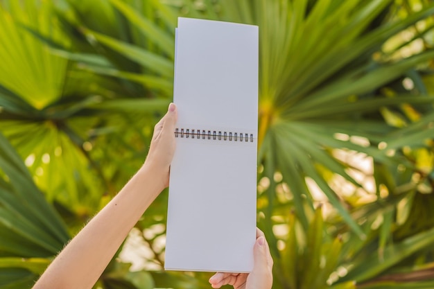 Manos de mujer en un fondo tropical sosteniendo una maqueta de papel de bloque de dibujo de letrero