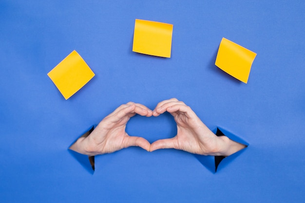 Manos de mujer en forma de corazón sobre un fondo azul. Las mujeres se insertan en un fondo de papel, en la parte superior hay tres palitos de papel. Lugar para una inscripción.