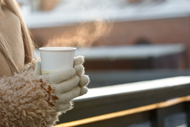 Manos de mujer en guantes blancos sosteniendo humeante taza blanca de café o té caliente en un día soleado de invierno frío