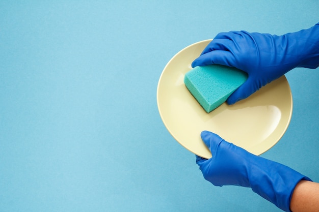 Manos de mujer en guantes protectores con placa beige y esponja sobre fondo azul. Concepto de lavado y limpieza.