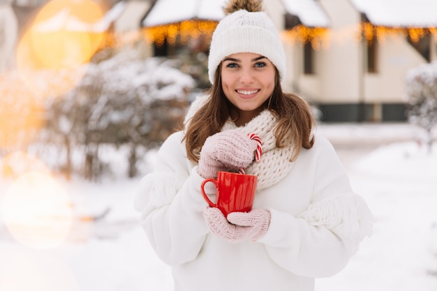 Manos de mujer en guantes que sostienen una taza acogedora con chocolate, té o café y un bastón de caramelo.