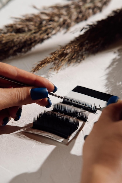 Las manos de una mujer irreconocible con manicura azul sosteniendo pinzas para la extensión de pestañas separando y tomando pestañas falsas de la paleta de cilios artificiales acostadas en la mesa cerca de la hierba de la pampa vertical