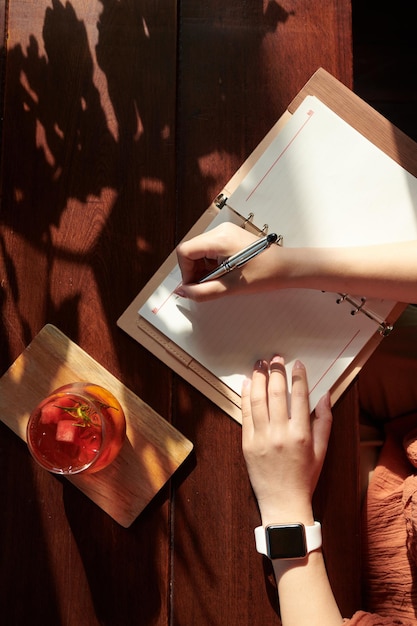 Manos de mujer joven creativa bebiendo cóctel de frutas y escribiendo en la vista del planificador desde la parte superior