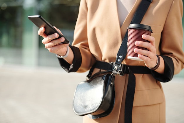 Manos de mujer joven elegante con bebida y bolso desplazándose a través de contactos en el teléfono inteligente mientras va a llamar a alguien al aire libre