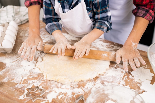 Manos de mujer joven y niño rodando masa sobre la mesa de la cocina mientras preparan galletas juntos