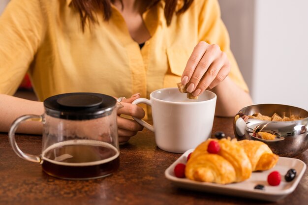 Manos de mujer joven poniendo dos cubos de azúcar morena en una taza con té o café mientras se va a desayunar por la mañana