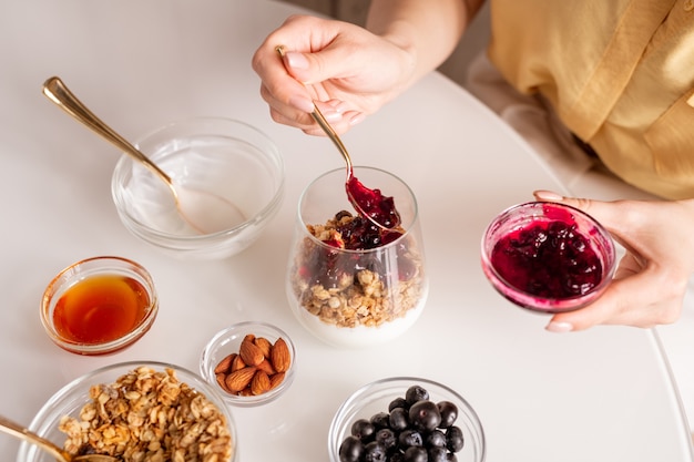 Manos de mujer joven poniendo mermelada de cereza apetitosa casera en vaso con muesli y crema agria mientras hace yogur para el desayuno