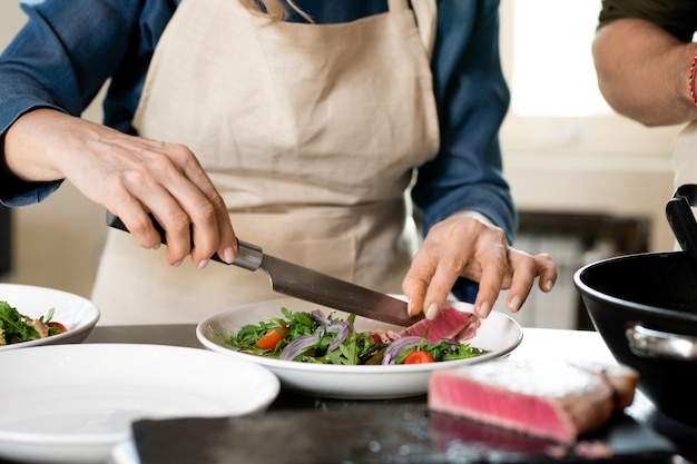 Manos de mujer madura poniendo una rodaja de carne ahumada en un plato con judías verdes al vapor con rúcula, cebolla picada y tomates cherry