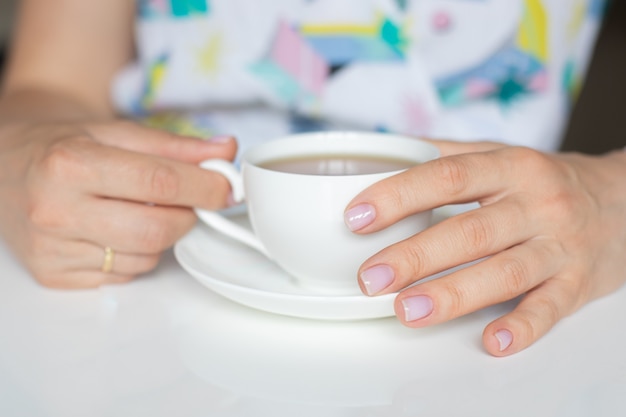 Manos de mujer con manicura de dedos de color nude con esmalte de uñas rosa sosteniendo una taza de té