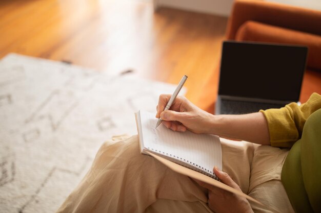 Las manos de una mujer mayor escribiendo en un cuaderno mientras está sentada junto a su portátil