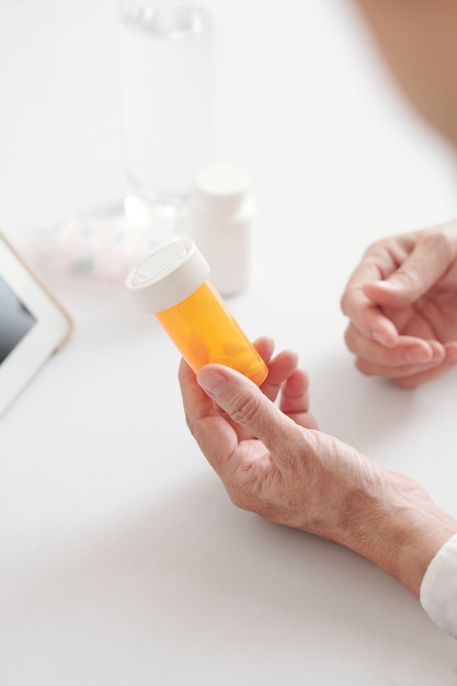 Manos de mujer mayor sosteniendo frasco de pastillas de plástico con analgésicos o suplementos