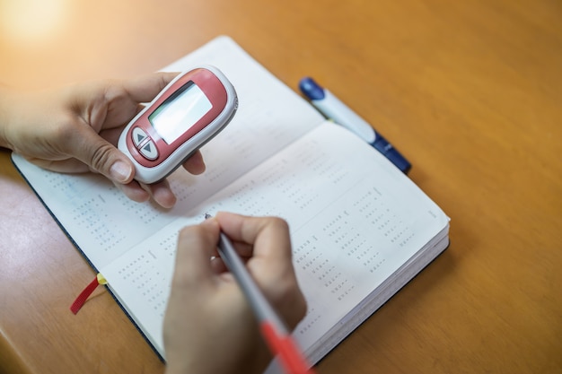 Las manos de la mujer que sostienen el medidor de glucosa y usan el bolígrafo para escribir el horario verifican el nivel de azúcar en la sangre