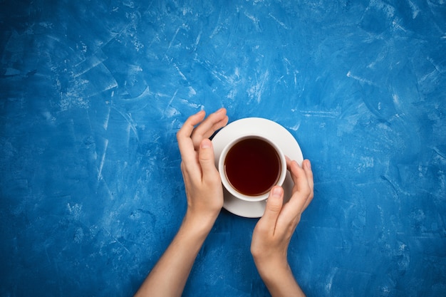 Manos de mujer sobre fondo de cemento azul con taza de té negro, flatlay