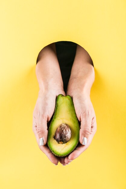 Manos de mujer sosteniendo un aguacate de un agujero negro en una pared de papel amarillo.