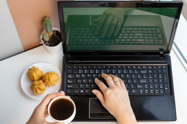 Manos de mujer sosteniendo café y deliciosos pasteles Mujer joven usando una computadora portátil mientras se relaja en casa con café