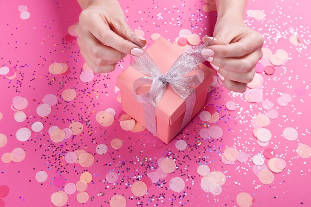 Manos de mujer sosteniendo caja de regalo con lazo plateado sobre fondo rosa pastel con brillo y confeti