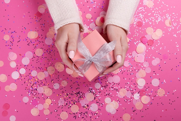 Manos de mujer sosteniendo caja de regalo con lazo plateado sobre fondo rosa pastel con brillo y confeti